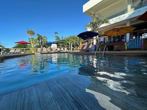 Uma piscina com espreguiçadeiras e guarda-sóis, cercada por palmeiras e um deck de madeira.