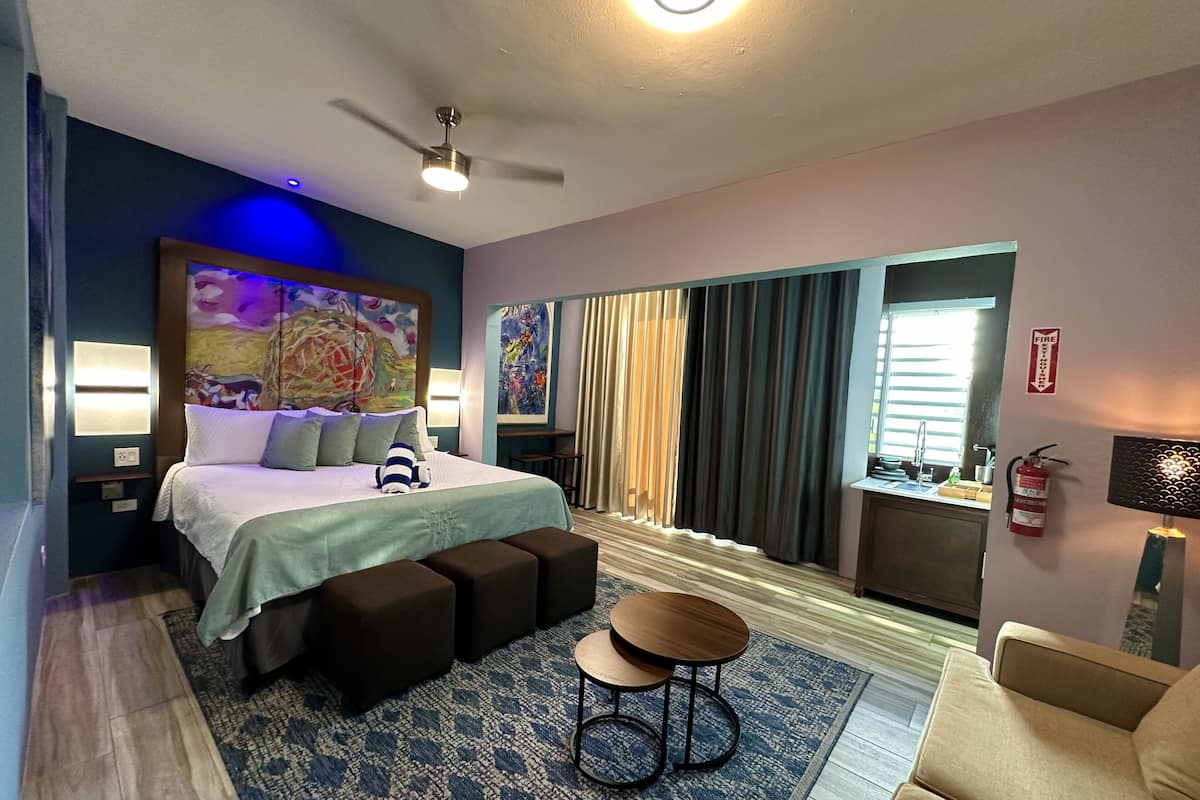 A modern hotel room with a large bed, a ceiling fan, a view of the outdoors, a fire extinguisher, and a small table.