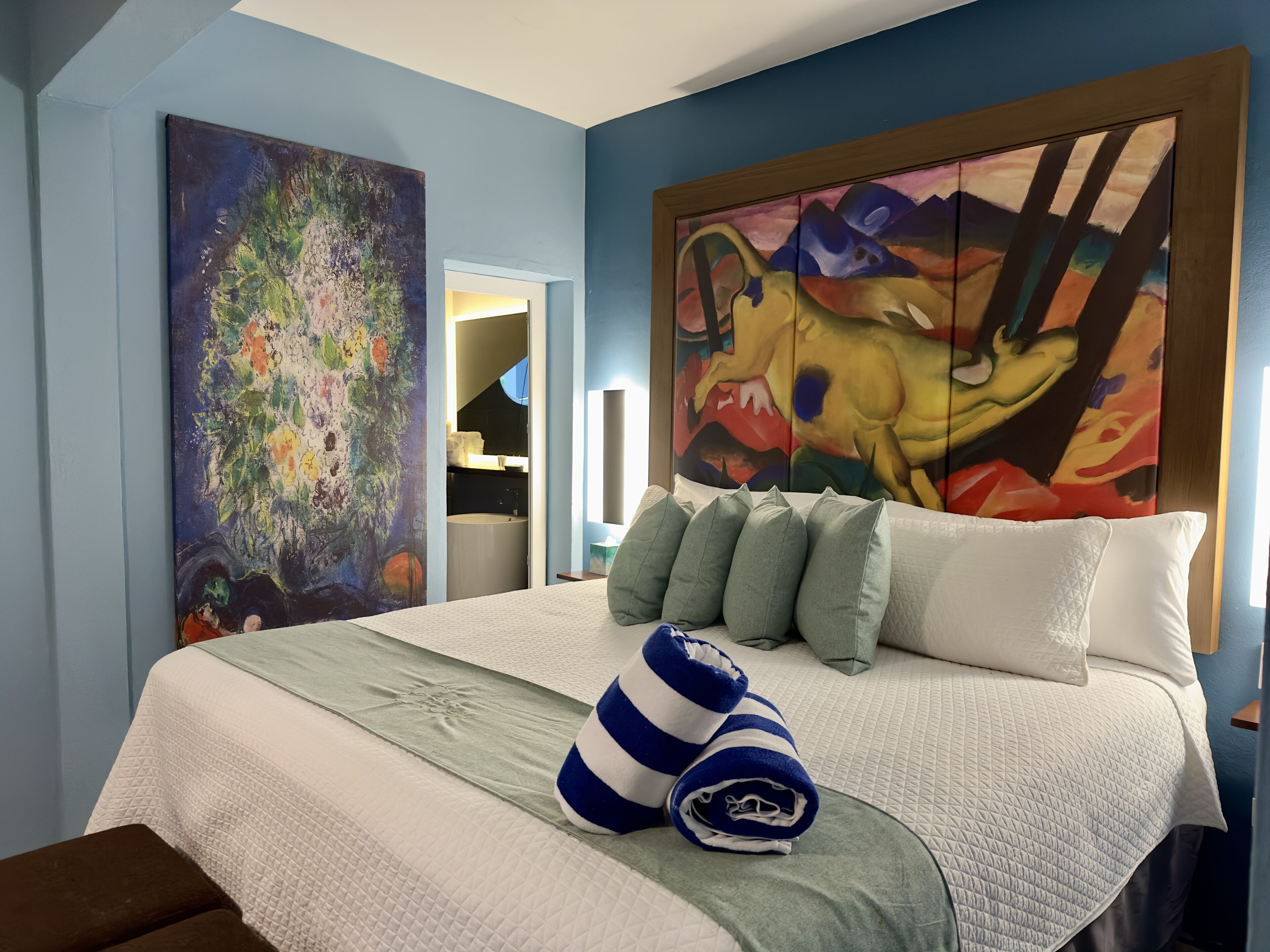 Habitación de hotel con una cama grande, un cuadro en la pared y un baño visible a través de una puerta abierta.