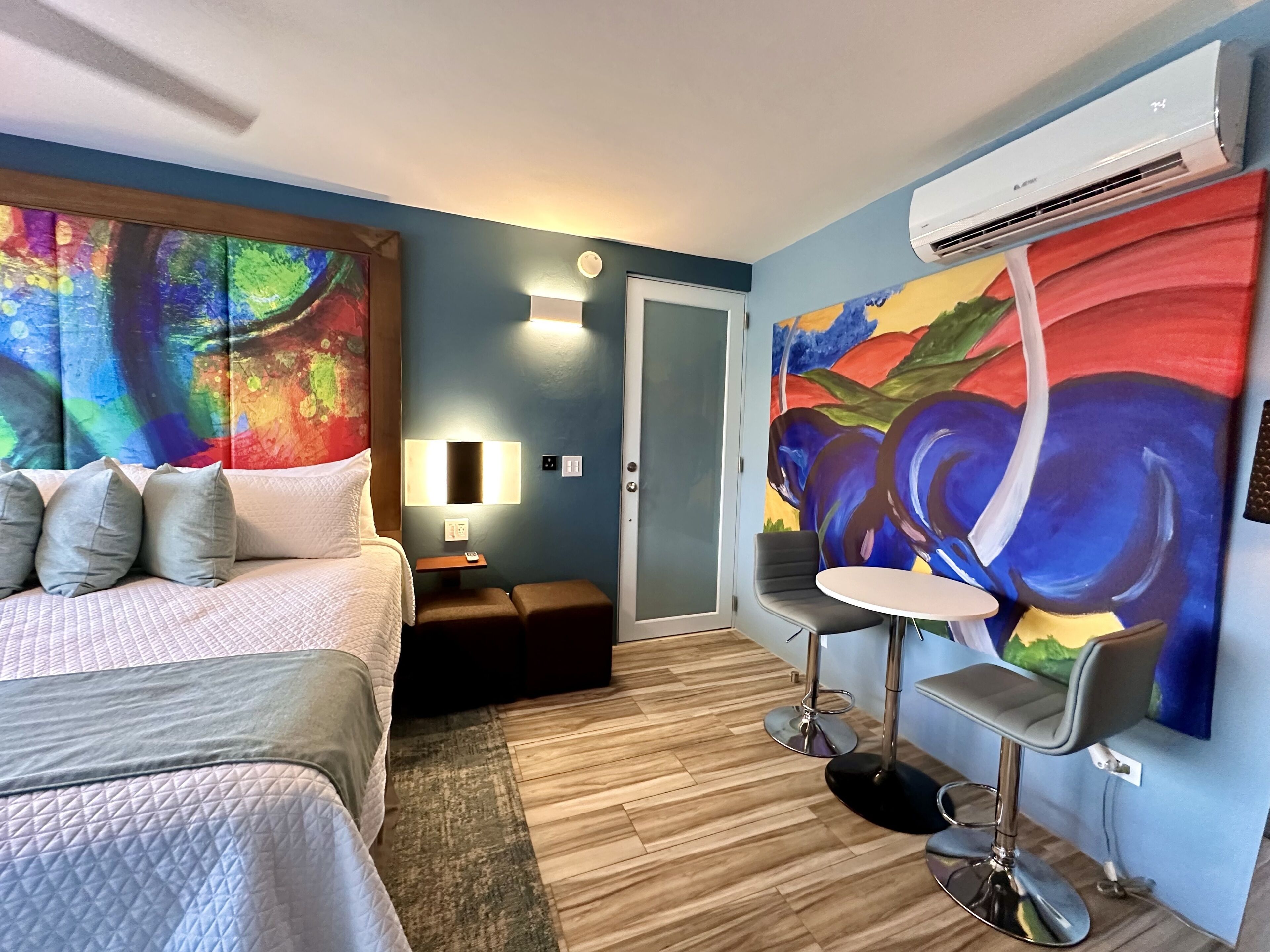 A hotel room with a bed, bedside table, lamp, two chairs, and a large abstract painting on the wall.