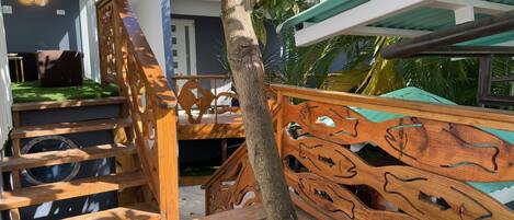 Een houten trap met ingewikkelde houtsnijwerken, die uitkomt op een balkon met uitzicht op een huis en omliggend groen.