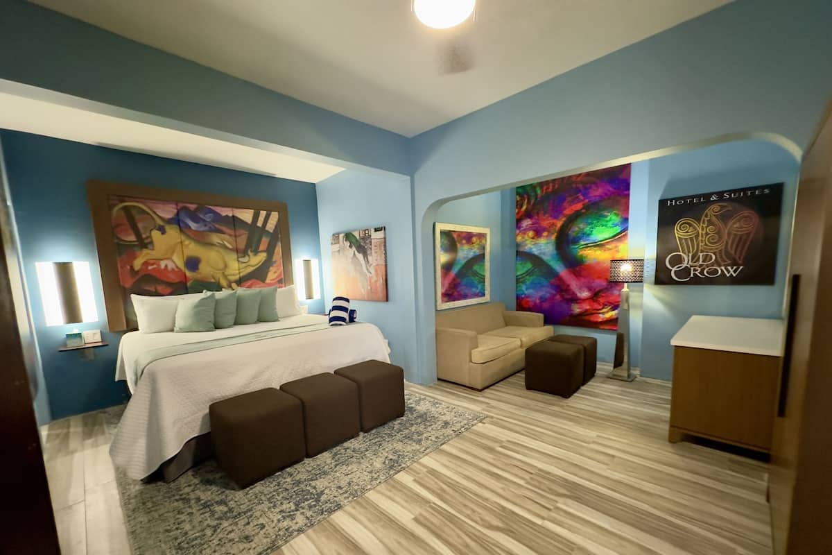 A modern bedroom with a bed, a sofa, and a desk. There are paintings on the walls and a lamp on the desk.