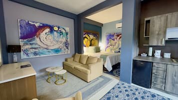 A modern hotel room with a sofa, a small round table, a bed with a nightstand, and a kitchenette area.