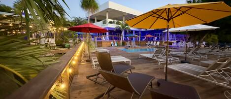 A poolside area with lounge chairs and umbrellas, a wooden deck, and string lights.