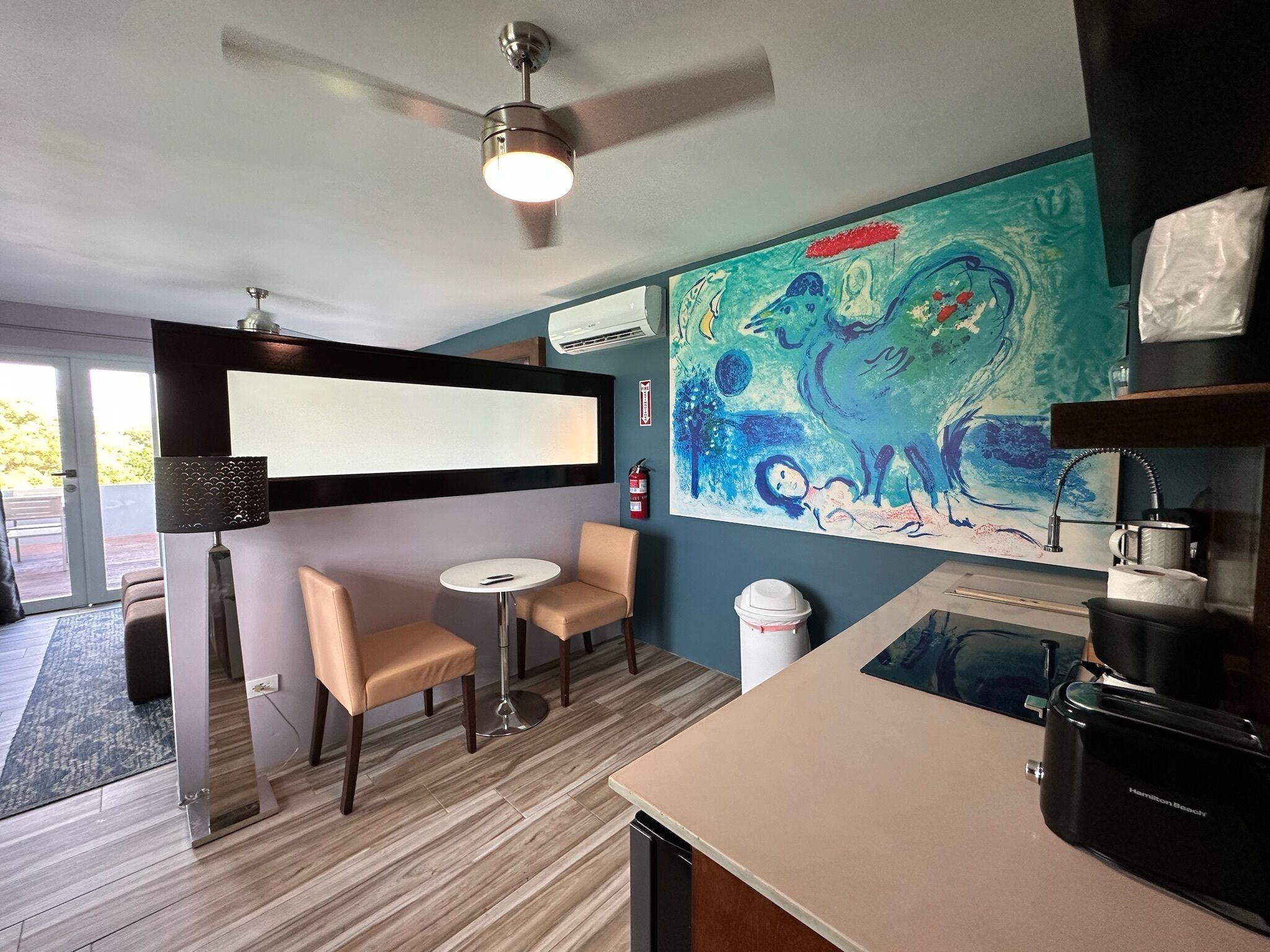 A modern room with a ceiling fan, a large abstract painting, a small round table with two chairs, a coffee machine, and a sink.