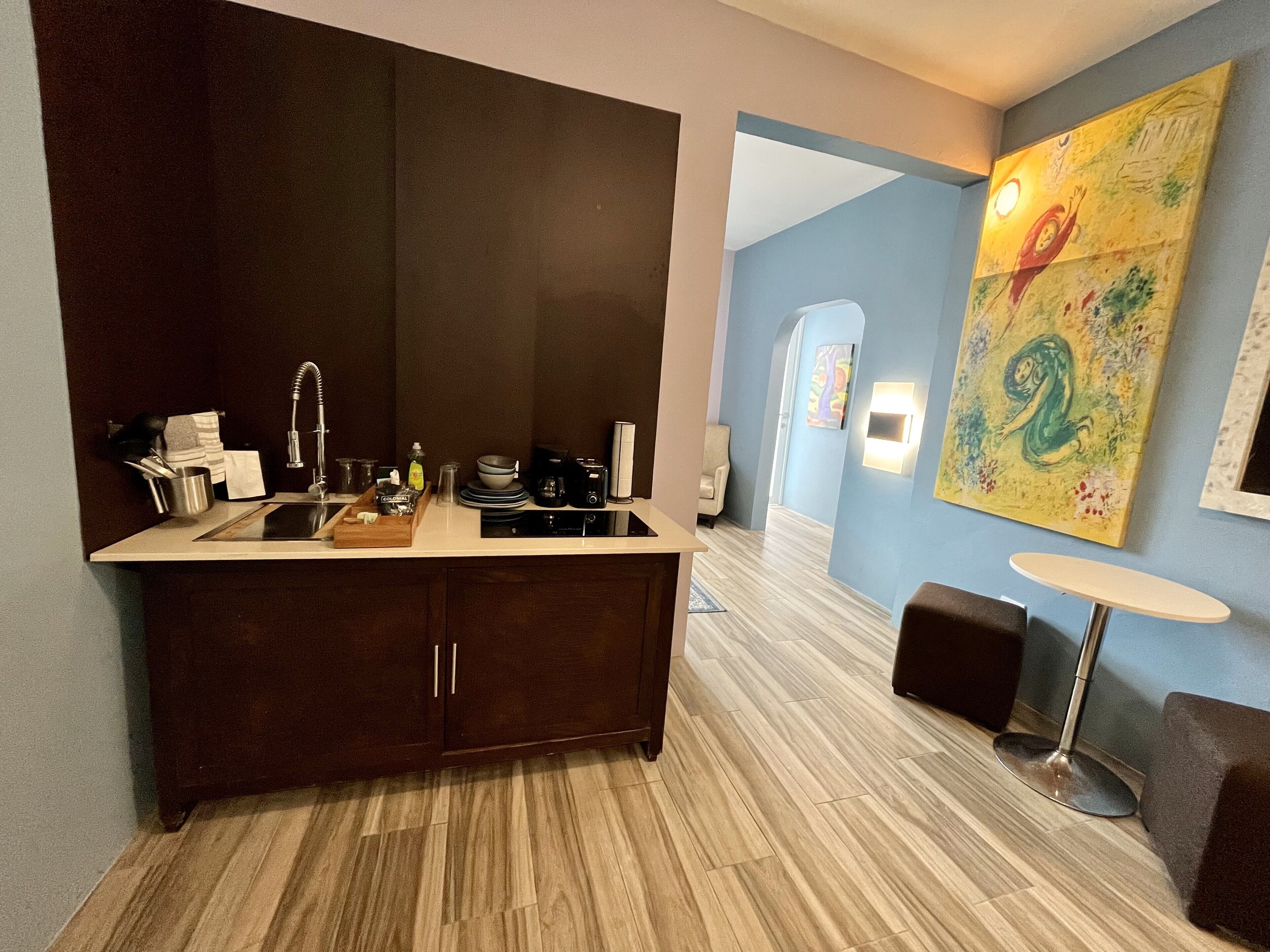 Una cocina moderna con armarios de madera oscura, electrodomésticos de acero inoxidable y una encimera de color claro. Hay un cuadro en la pared y una pequeña mesa redonda con una lámpara.