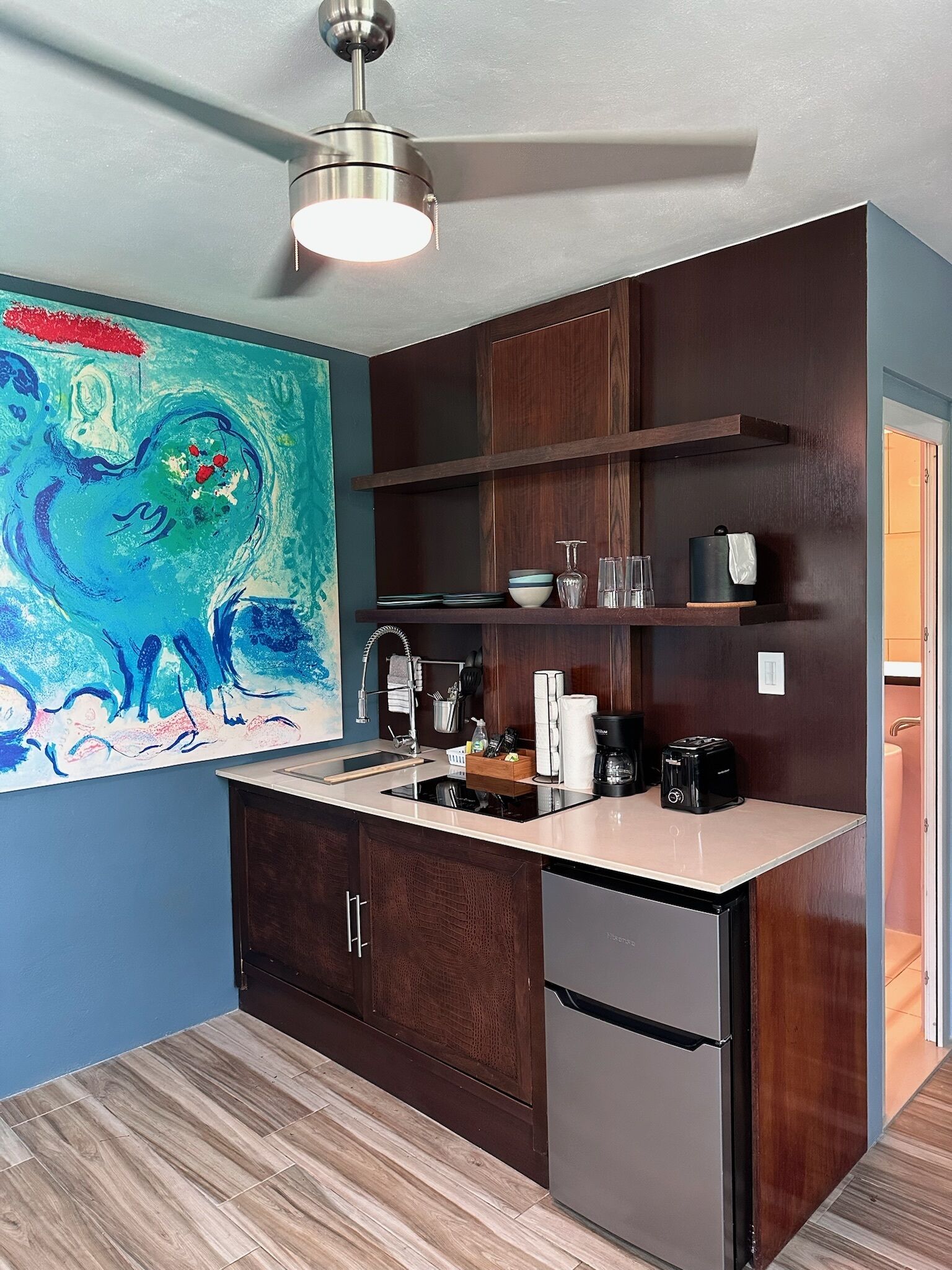 A compact kitchen with dark wood cabinets, a stainless steel refrigerator, a built-in oven, and a small countertop. There is a ceiling light fixture and a colorful abstract painting on the wall.