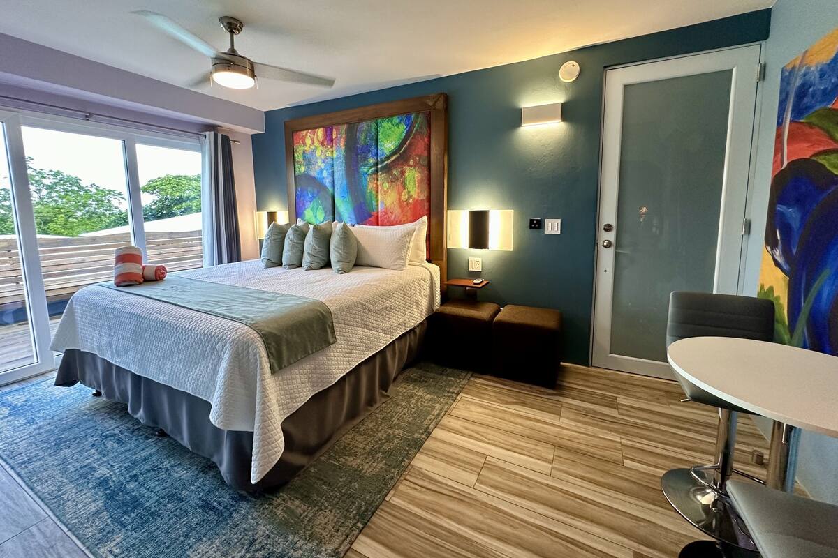 A modern hotel room with a large bed, a desk, and a chair. There is a window with a view of greenery outside.