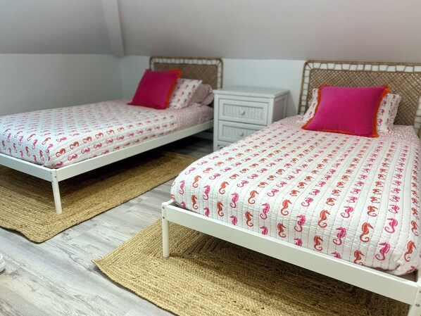Dos camas individuales con estructura blanca y ropa de cama estampada, cada una con un almohadón rosa, sobre un piso de madera con alfombras tejidas.