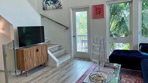 A modern living room with a staircase, a TV on a wooden cabinet, a glass coffee table with decorative items, and large windows with a view of greenery.