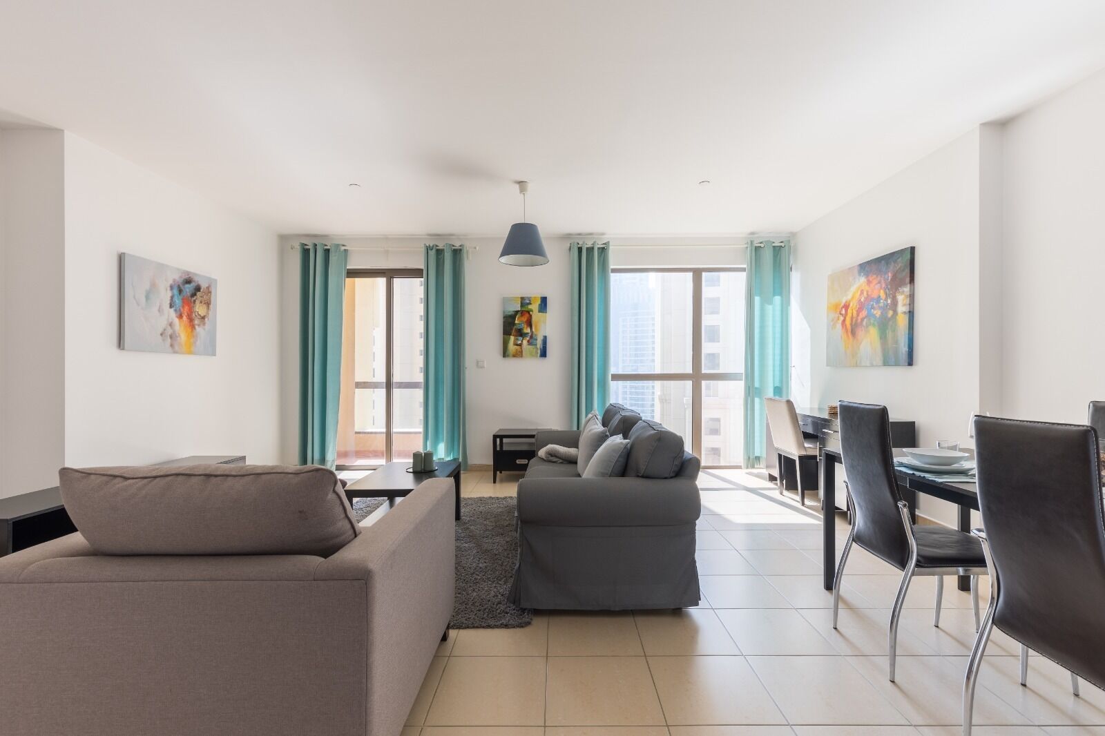 Un salon moderne avec un canapé gris, un fauteuil gris et un coin repas comprenant une table et des chaises. Il y a deux fenêtres avec des rideaux de couleur turquoise, un luminaire au plafond et des œuvres d’art accrochées aux murs.