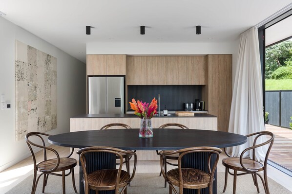 A modern kitchen with wooden cabinets, a stainless steel refrigerator, and a dining area with a round table and wicker chairs. - Black House Onetangi Beach | You've Got It Maid (Waiheke Island)
