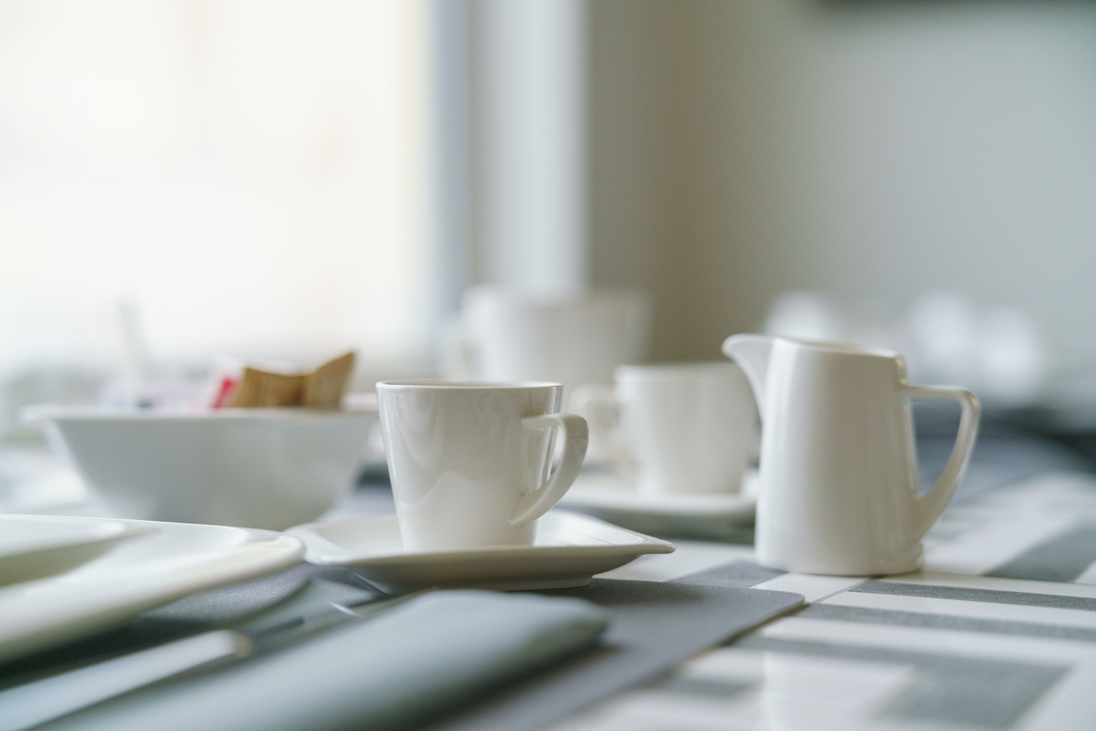 A table with white ceramic cups and a pitcher, set on a patterned tablecloth.