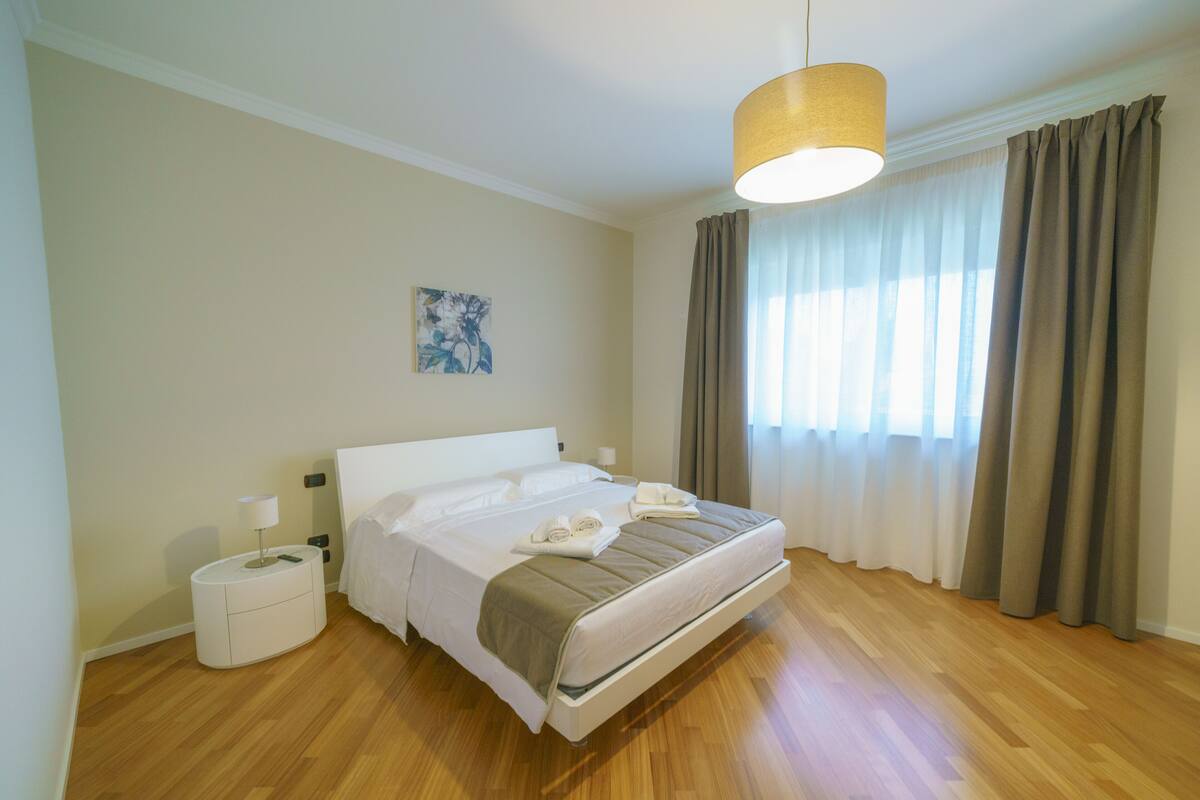 Una habitación de hotel con cama, mesitas de noche, lámpara, un cuadro en la pared y ventana con cortinas.