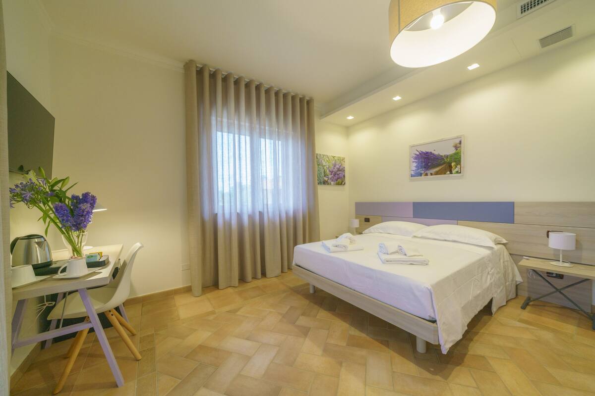 Habitación de hotel con cama, escritorio con silla, mesita de noche con lámpara y ventana con cortinas.