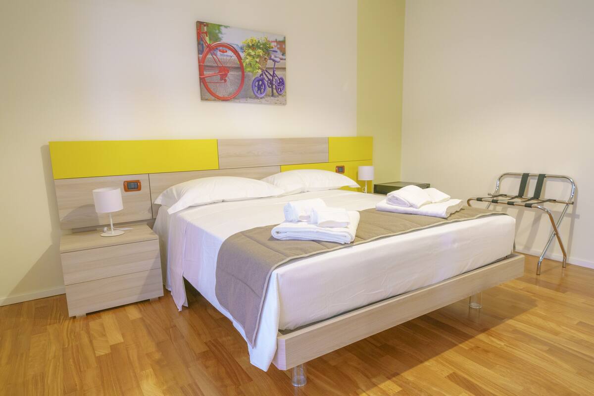 Una habitación de hotel moderna con suelo de madera, una cama con ropa blanca, una mesita de noche con lámpara y una obra de arte con temática de bicicletas en la pared.