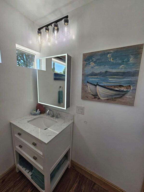 A bathroom with a white vanity, a large mirror, a painting of a boat on water, and a skylight.