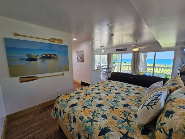A bedroom with a large bed, a painting of boats on the wall, and a view of the ocean through the window.