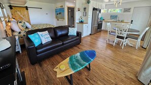 Un salon avec un canapé en cuir noir, une table basse en bois ornée d’un motif de planche de surf, un coin repas avec des chaises blanches et, au fond, une cuisine.