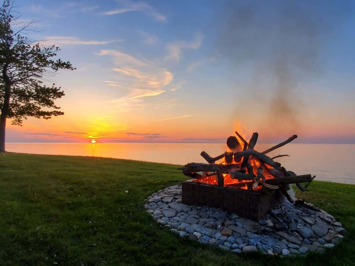 A campfire at sunset with a view of the water and a tree in the background.