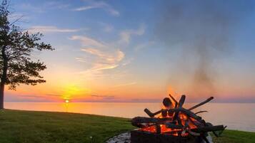 A campfire at sunset with a view of the water and a tree in the background.