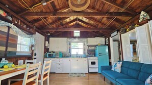 A cozy cabin interior with a wooden ceiling, a dining area with a table and chairs, a kitchenette with a blue refrigerator, and a blue sofa.