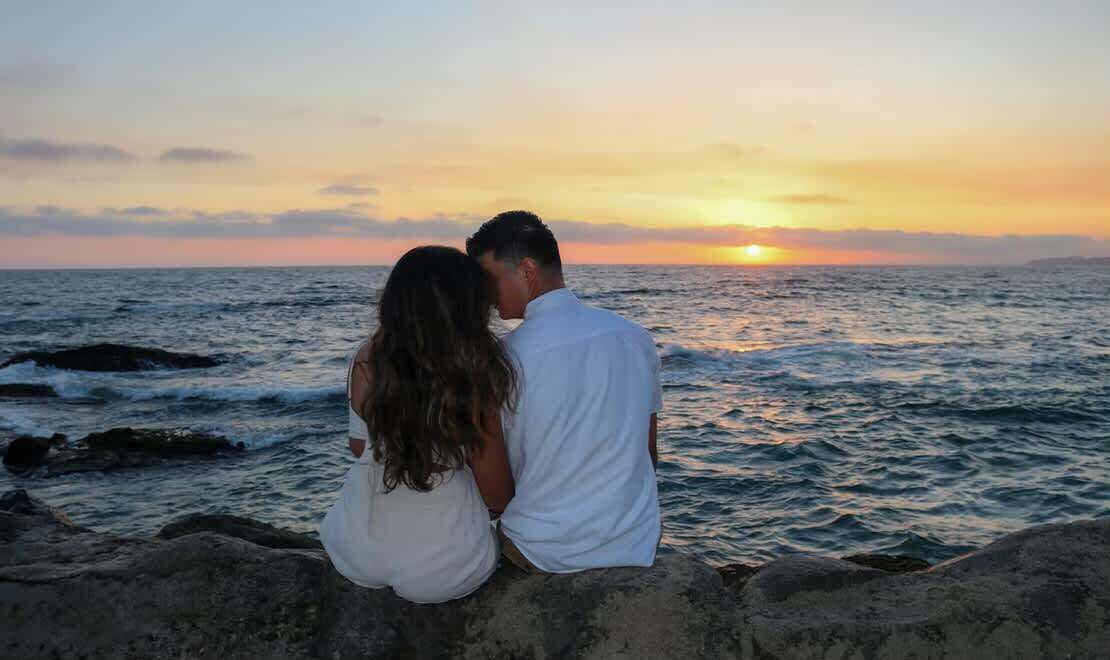 A couple sitting on a rocky ledge overlooking the ocean at sunset.
