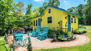 A yellow house with a blue roof, surrounded by a brick patio, blue chairs, and a small garden.