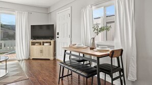 Una sala de estar moderna con una mesa de comedor de madera, sillas de cuero negro, un televisor de pantalla plana y un amplio ventanal con cortinas blancas.