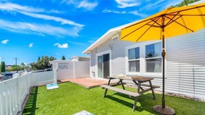 Patio trasero con una mesa de picnic y sombrilla, rodeado por una cerca blanca y una casa con ventanas.