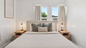Un dormitorio con una cama grande, dos mesitas de noche, una ventana con cortinas e imagen enmarcada en la pared.