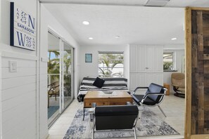 A room with a bed, a wooden chest, a black chair, and a window with a view of palm trees.