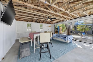 A room with a wooden ceiling, a television mounted on the wall, a sofa, a table with chairs, and a window with a view of palm trees.