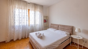 Un dormitorio con una cama, una mesita de noche, una lámpara y una ventana con cortinas traslúcidas.