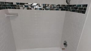 Une baignoire blanche dotée d’un pommeau de douche, entourée de carreaux de céramique blancs rectangulaires et ornée d’une bordure décorative en carreaux.