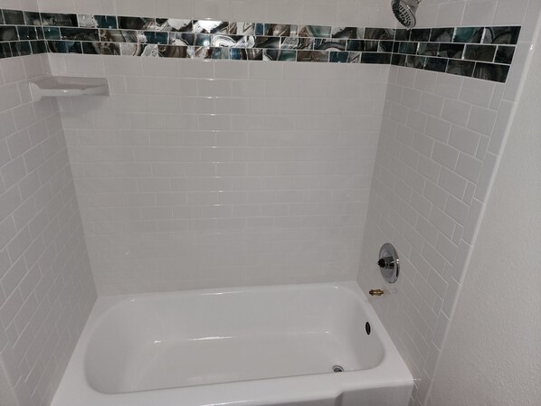 Una bañera blanca con cabezal de ducha, rodeada de azulejos blancos tipo metro y un borde decorativo de azulejos.