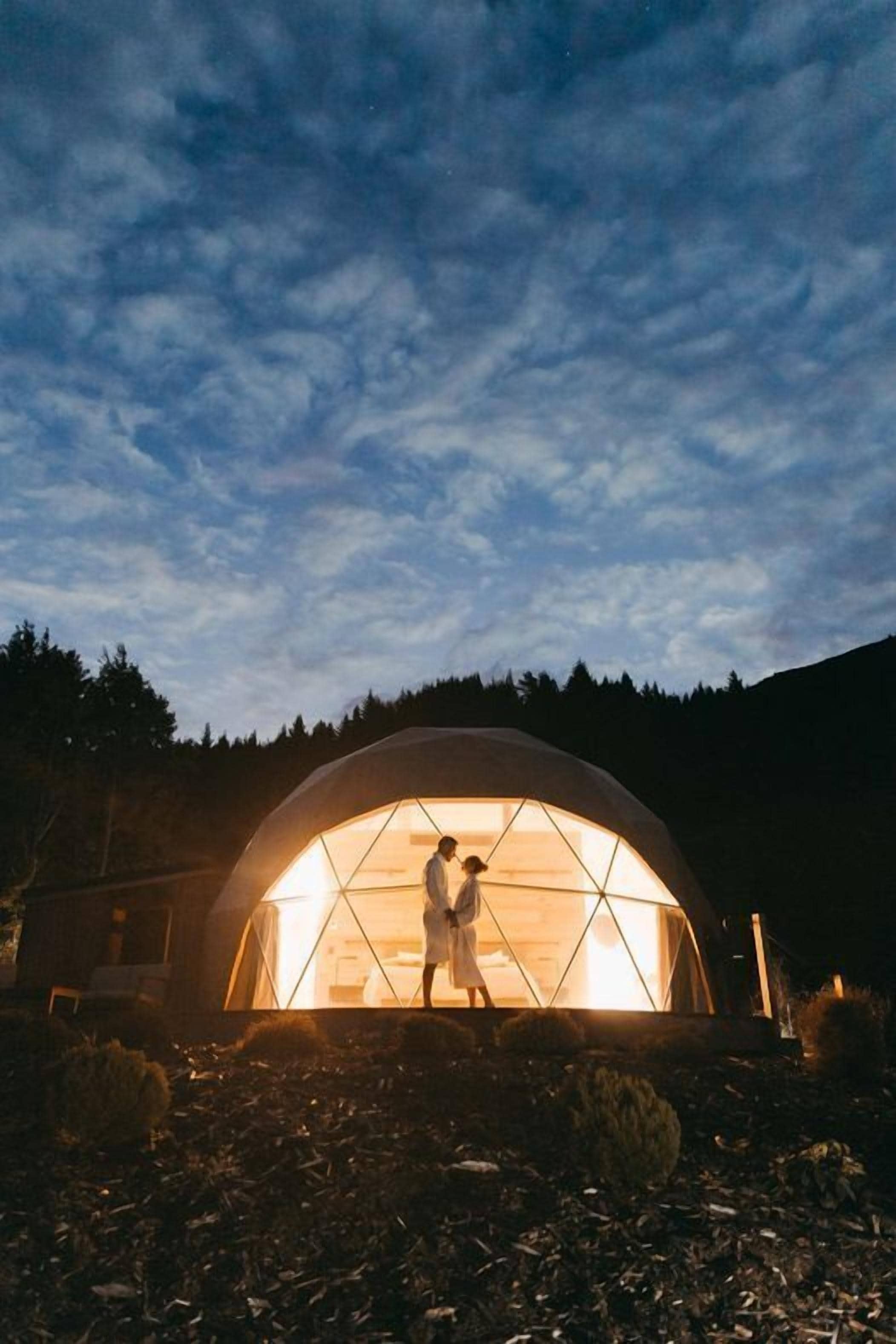 A geodesic dome structure with a warm interior light, surrounded by a forested landscape.