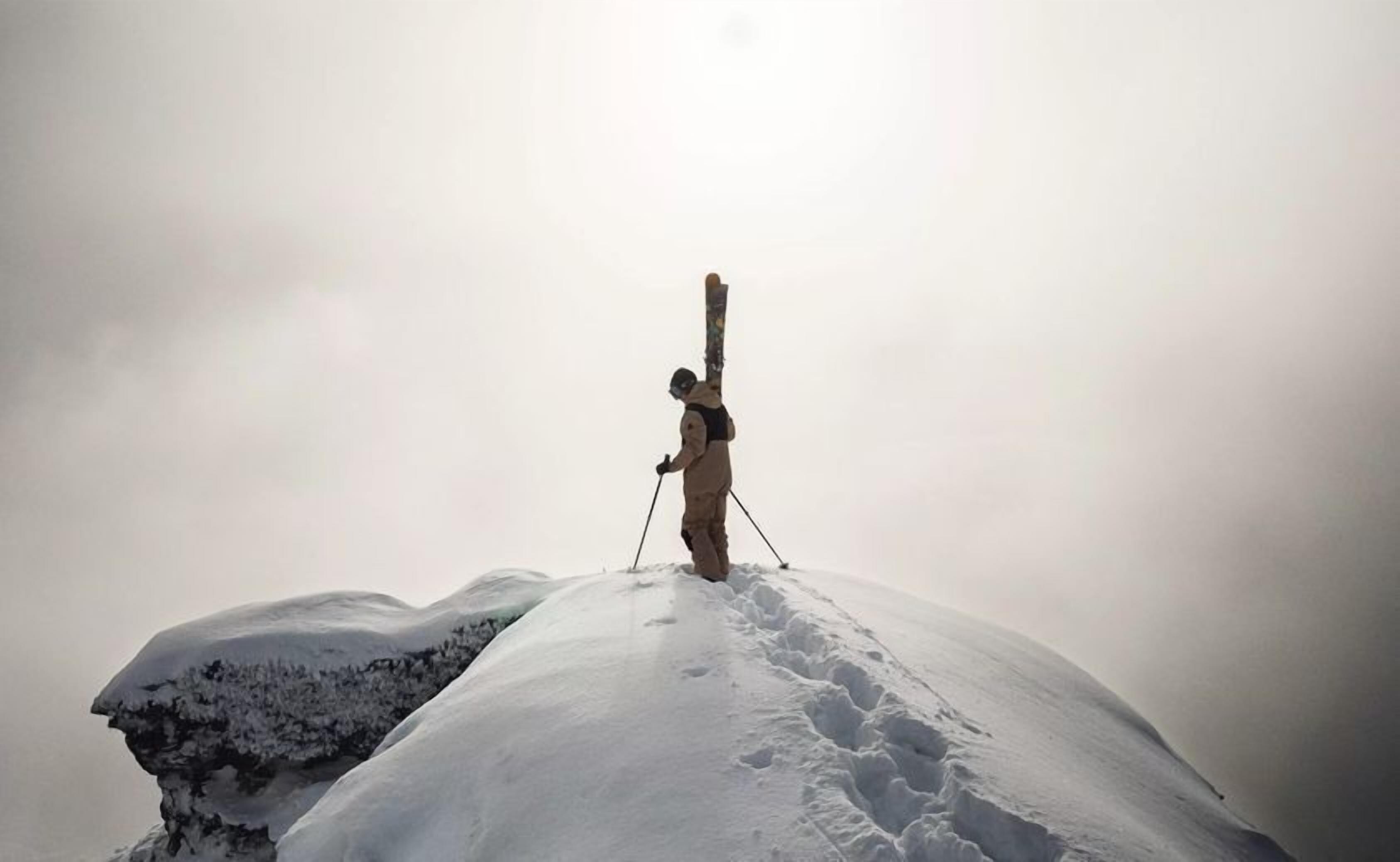 A person standing on a snow-covered peak with skis.