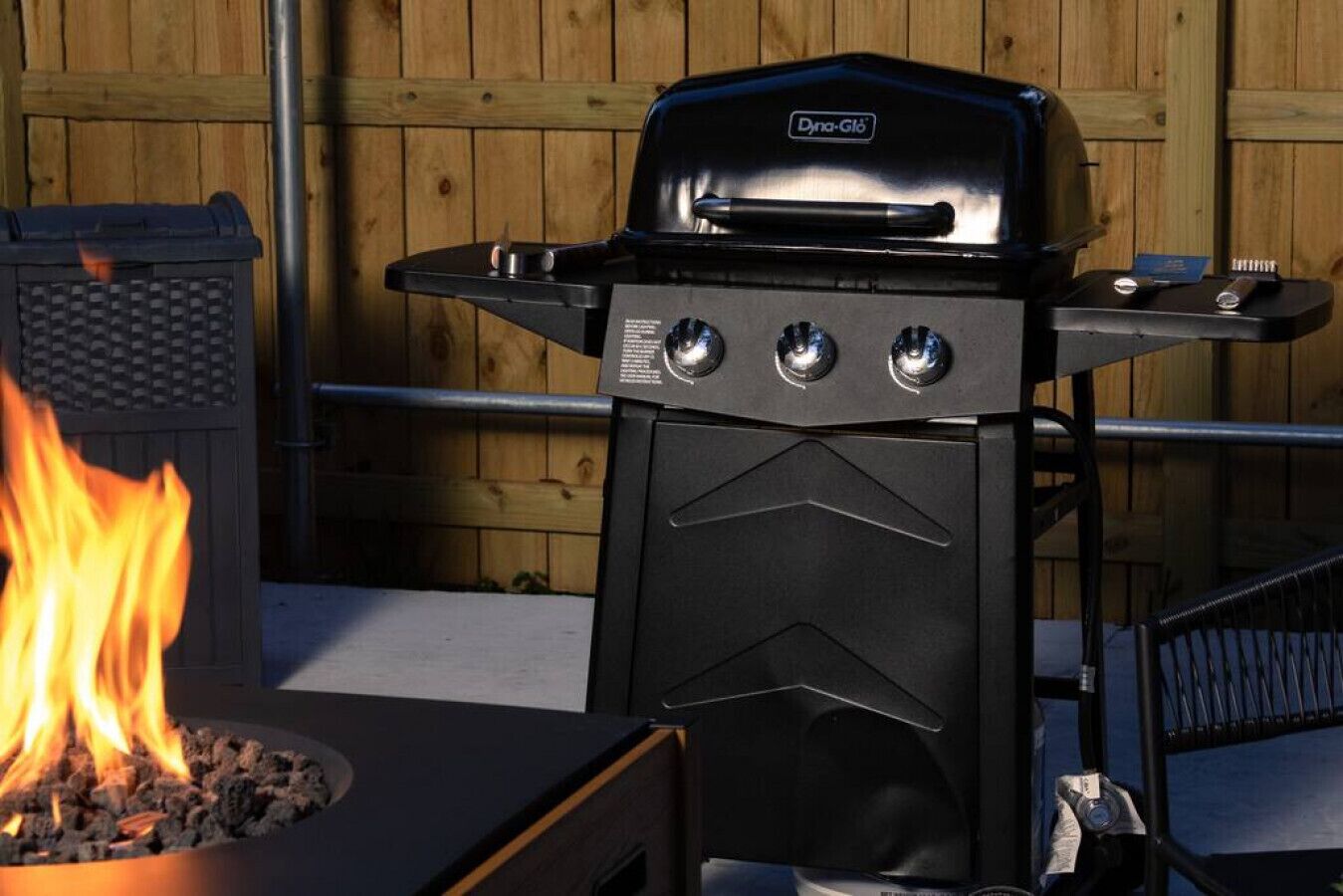 A black outdoor grill with three control knobs and a side burner, placed against a wooden fence backdrop.