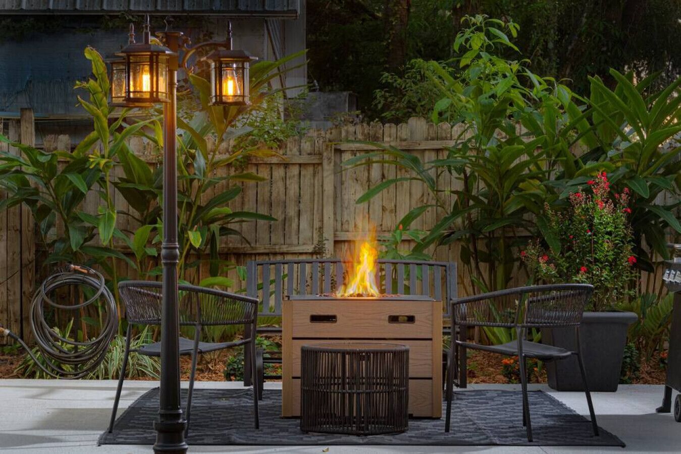 Een terras buiten met een houten vuurkorf, twee metalen stoelen en twee lantaarnlampen.