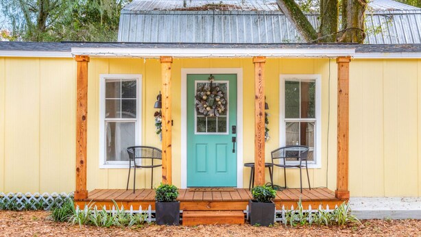 Uma casa amarela com porta verde, duas janelas e varanda com duas cadeiras.