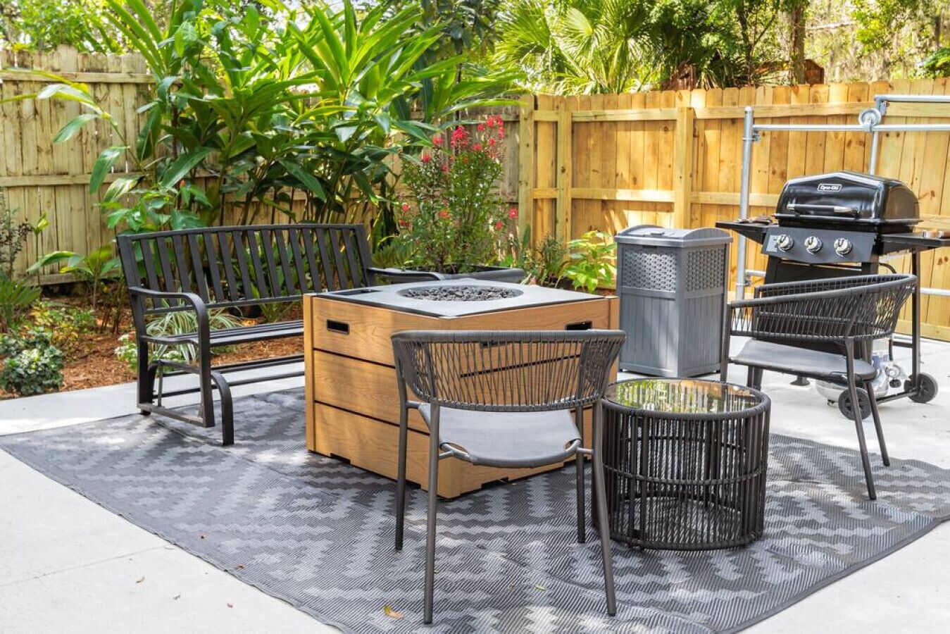 Un patio al aire libre con una mesa de madera, dos sillas, un basurero y una parrilla.