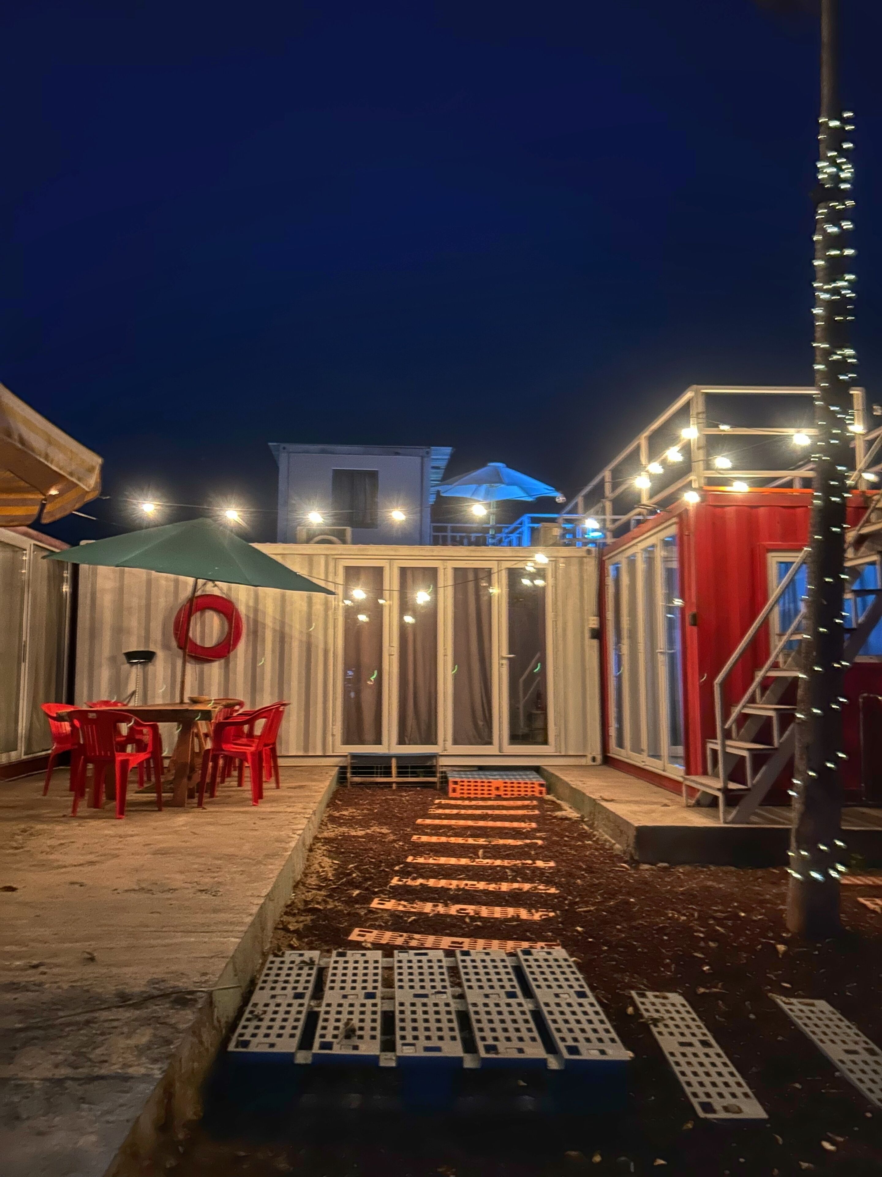 An outdoor patio area with a table and chairs, a covered seating area, and a pathway leading to a building entrance.