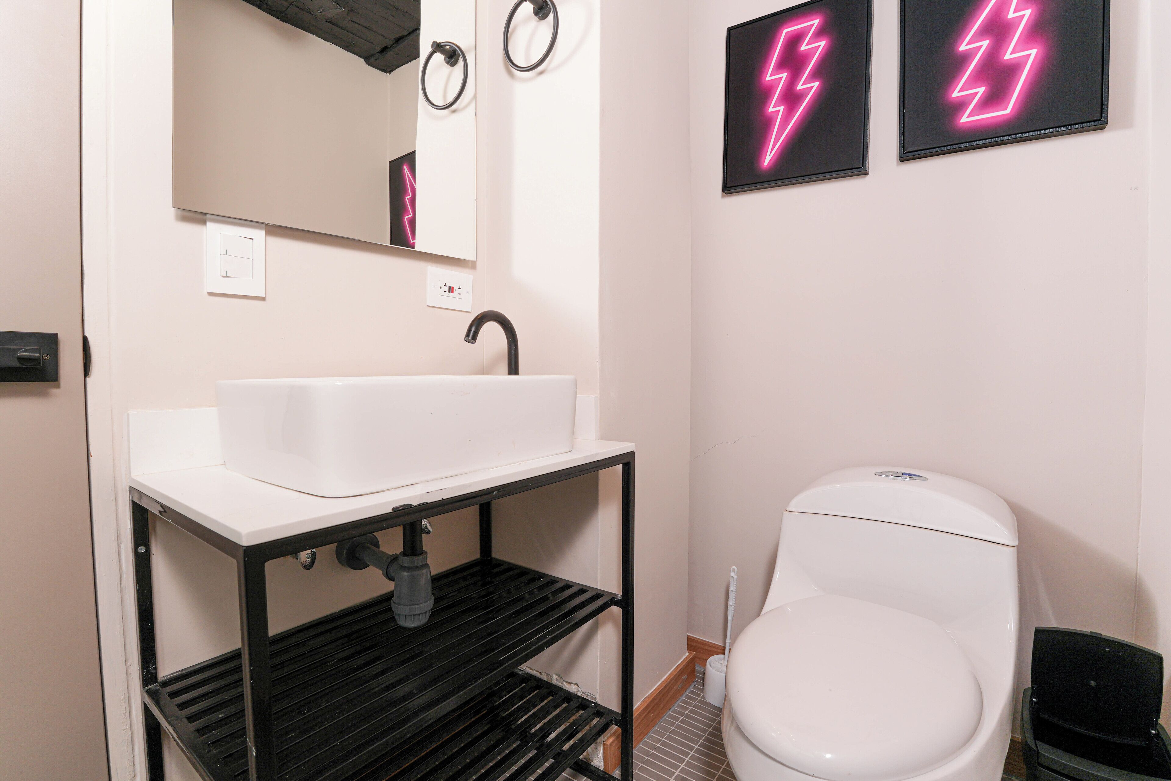 Une salle de bain avec un lavabo blanc, des étagères noires, un miroir et deux enseignes néon en forme d’éclair sur le mur.