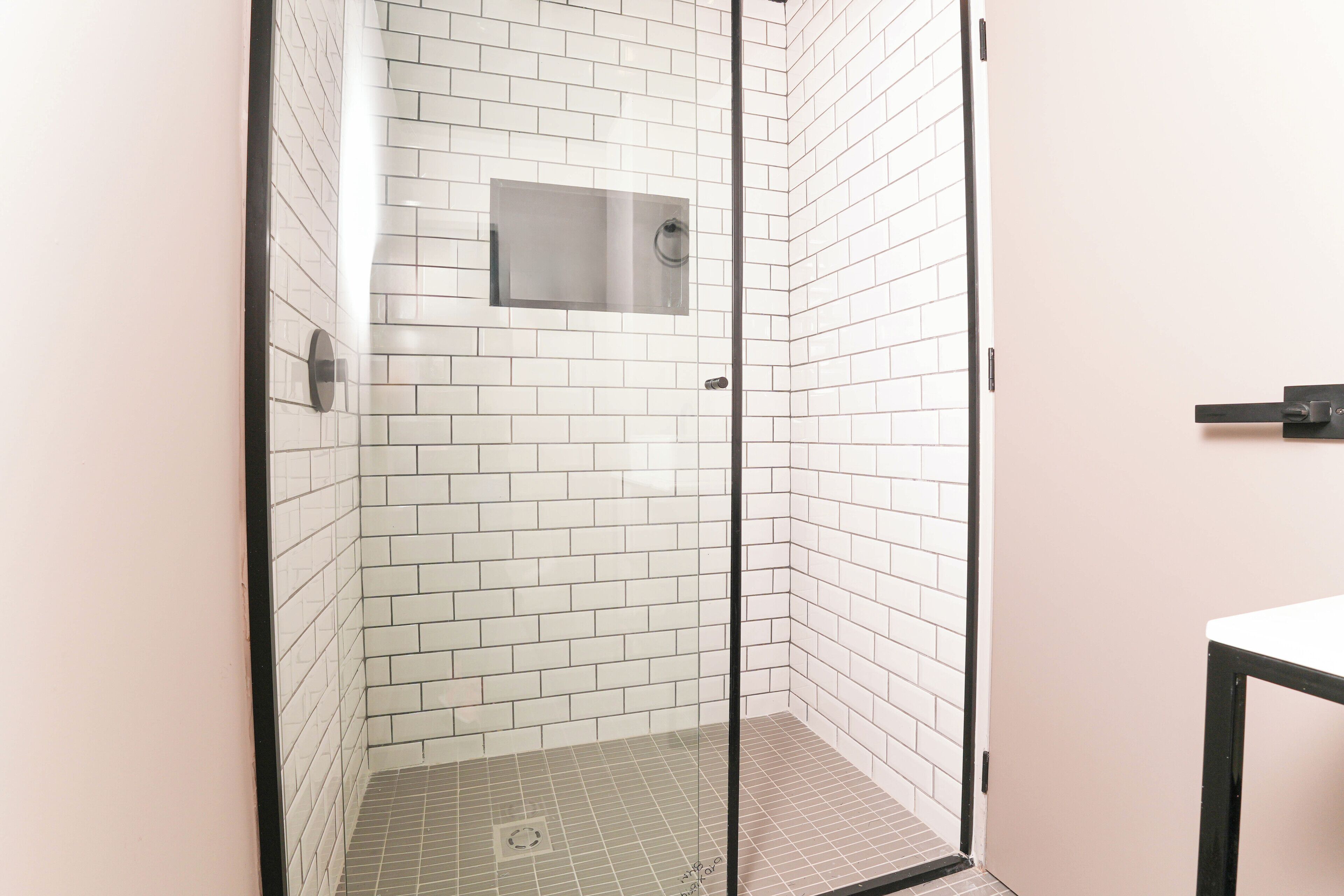 Ducha en esquina con paredes y suelo de azulejos blancos, puerta de vidrio y un espejo pequeño en la pared.