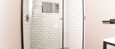 Ducha en esquina con paredes y suelo de azulejos blancos, puerta de vidrio y un espejo pequeño en la pared.
