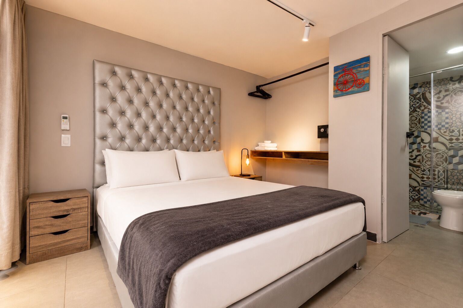 Une chambre à coucher avec une tête de lit capitonnée, une table de chevet avec une lampe, une commode en bois et une salle de bain carrelée avec des motifs.