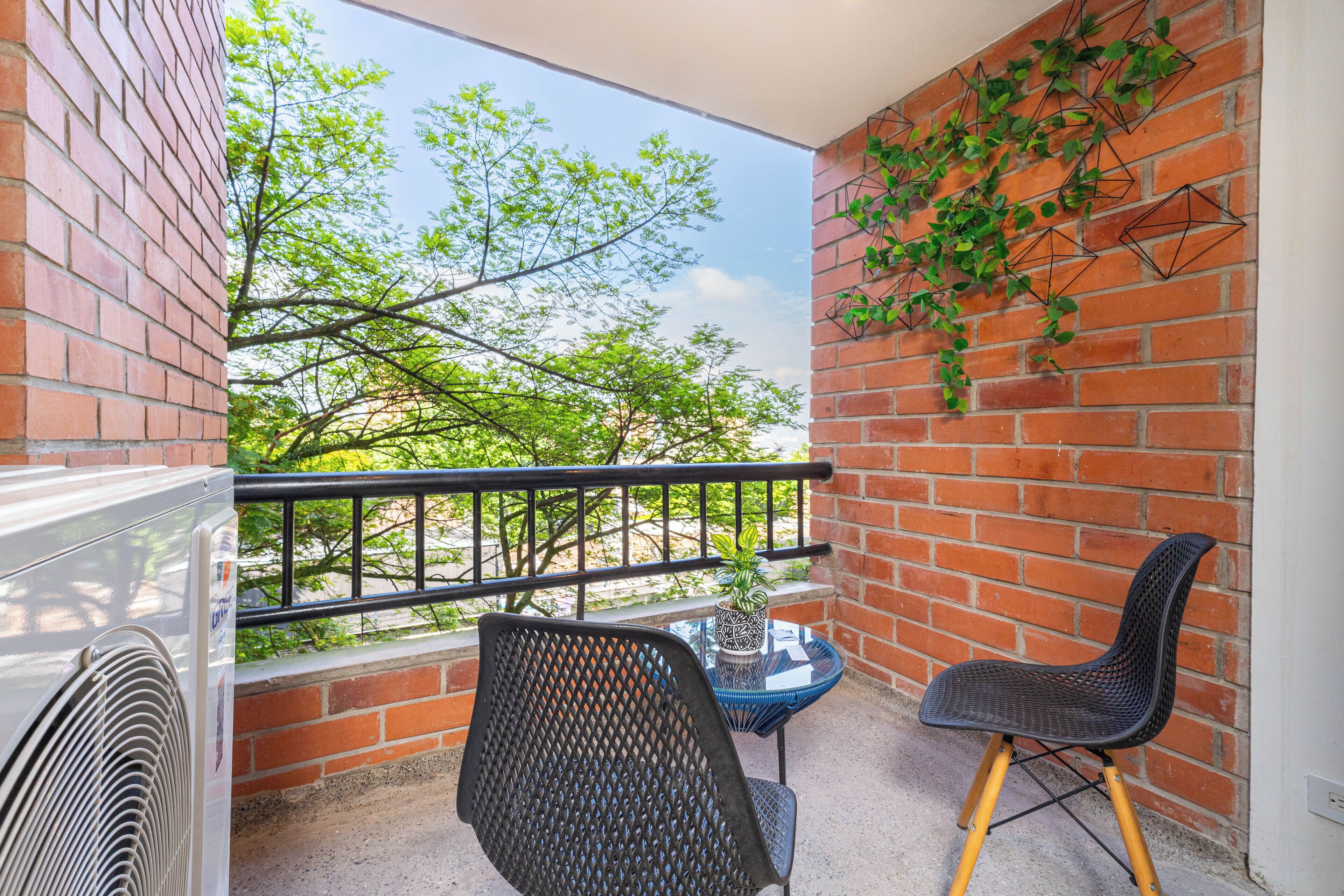 A balcony with a brick wall, a black chair, a small table with a plant, and a fan.