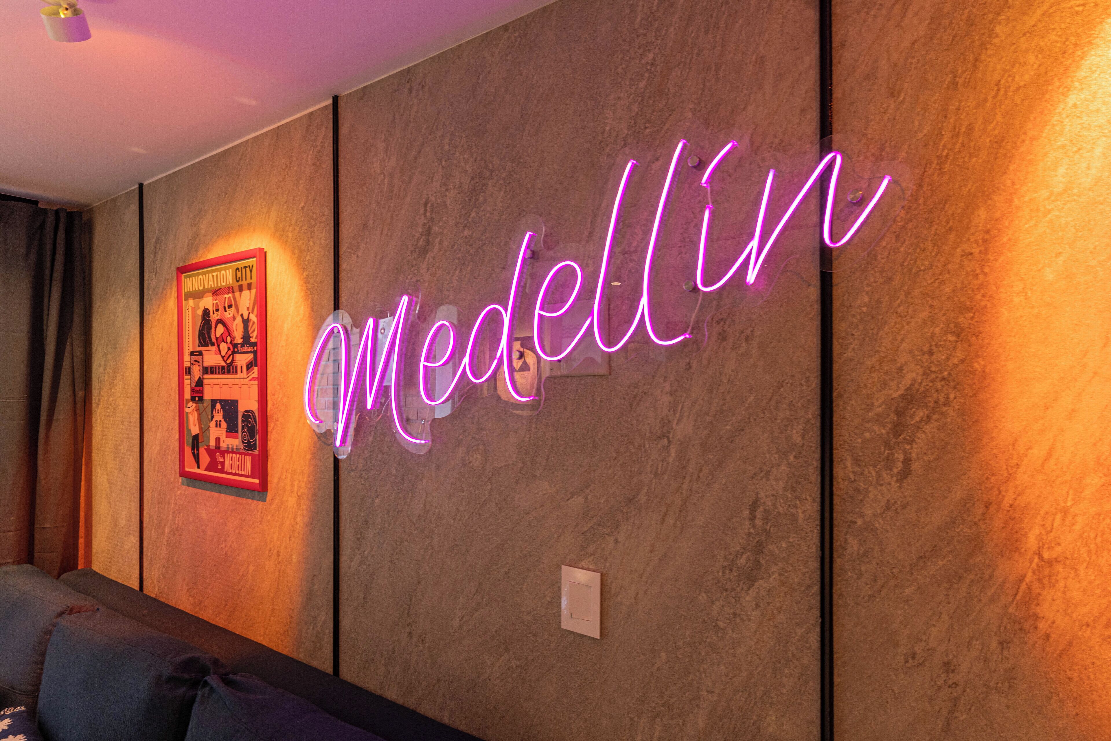 A neon sign spelling "Medellin" on a textured wall, a framed poster, and a light switch.