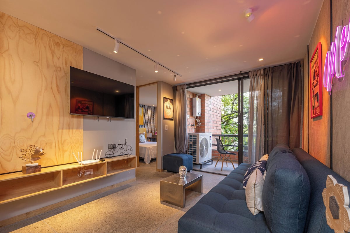 A modern living room with a sofa, a coffee table, and a TV mounted on the wall. There is a balcony with a view of greenery outside.